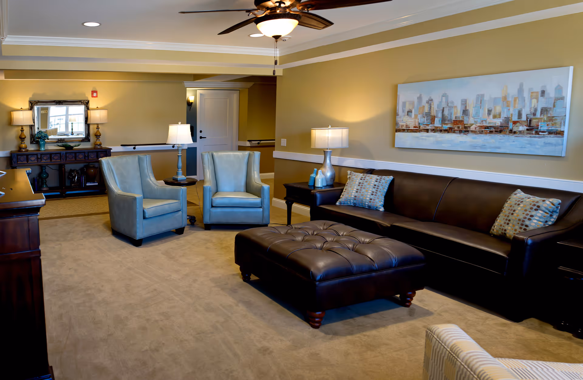 A cozy living room area with a dark brown leather sofa adorned with two patterned pillows, a large tufted leather ottoman in front, and two light blue armchairs. The walls are painted beige with white trim, and a large painting of a city skyline hangs above the sofa. The room is softly lit by table lamps on dark wood side tables and a ceiling fan with a light fixture. A wooden console table with decorative items and a mirror is visible in the background.