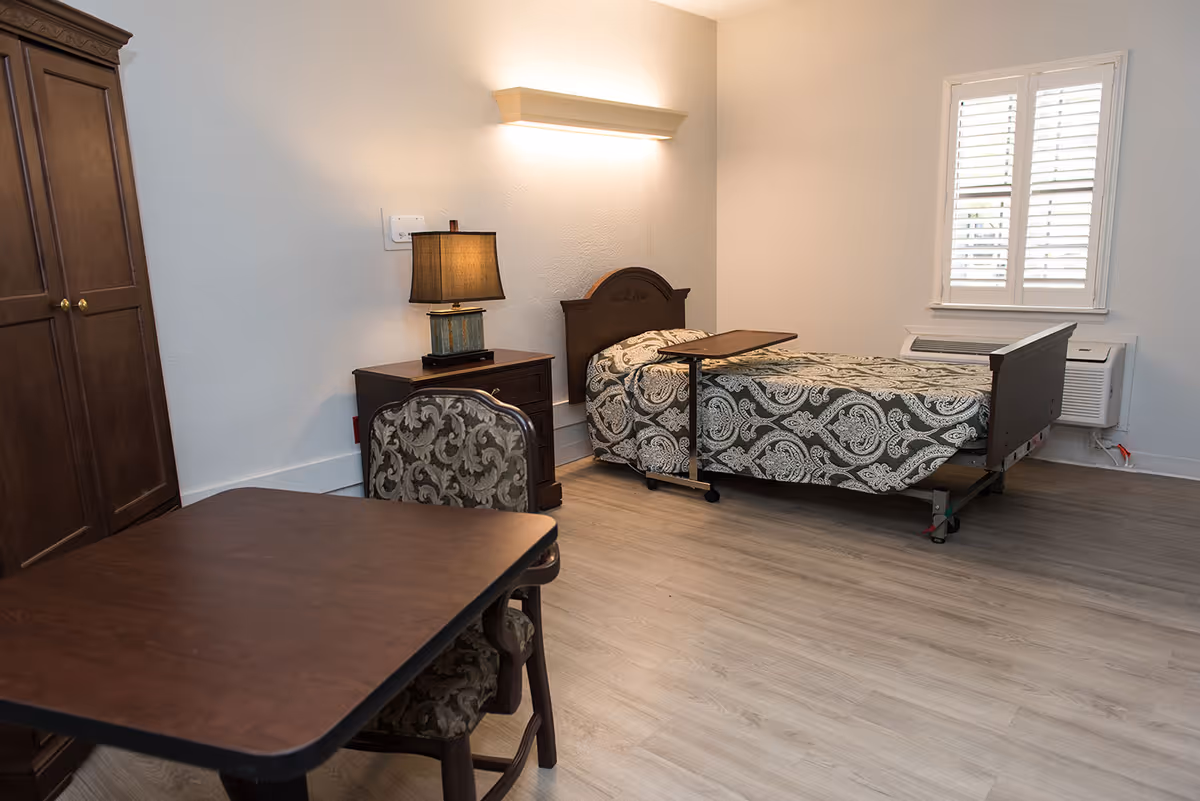 A simple, clean bedroom in a senior living facility with a single bed covered in a patterned bedspread, a wooden nightstand with a lamp, a wooden wardrobe, a table with two upholstered chairs, and a window with white shutters. The room has light-colored wooden flooring and white walls.