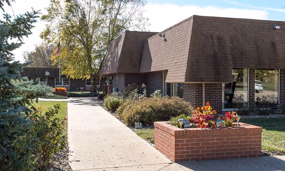 Exterior front of a brick building with a sidewalk, raised brick planter with flowers, and an American flag.
