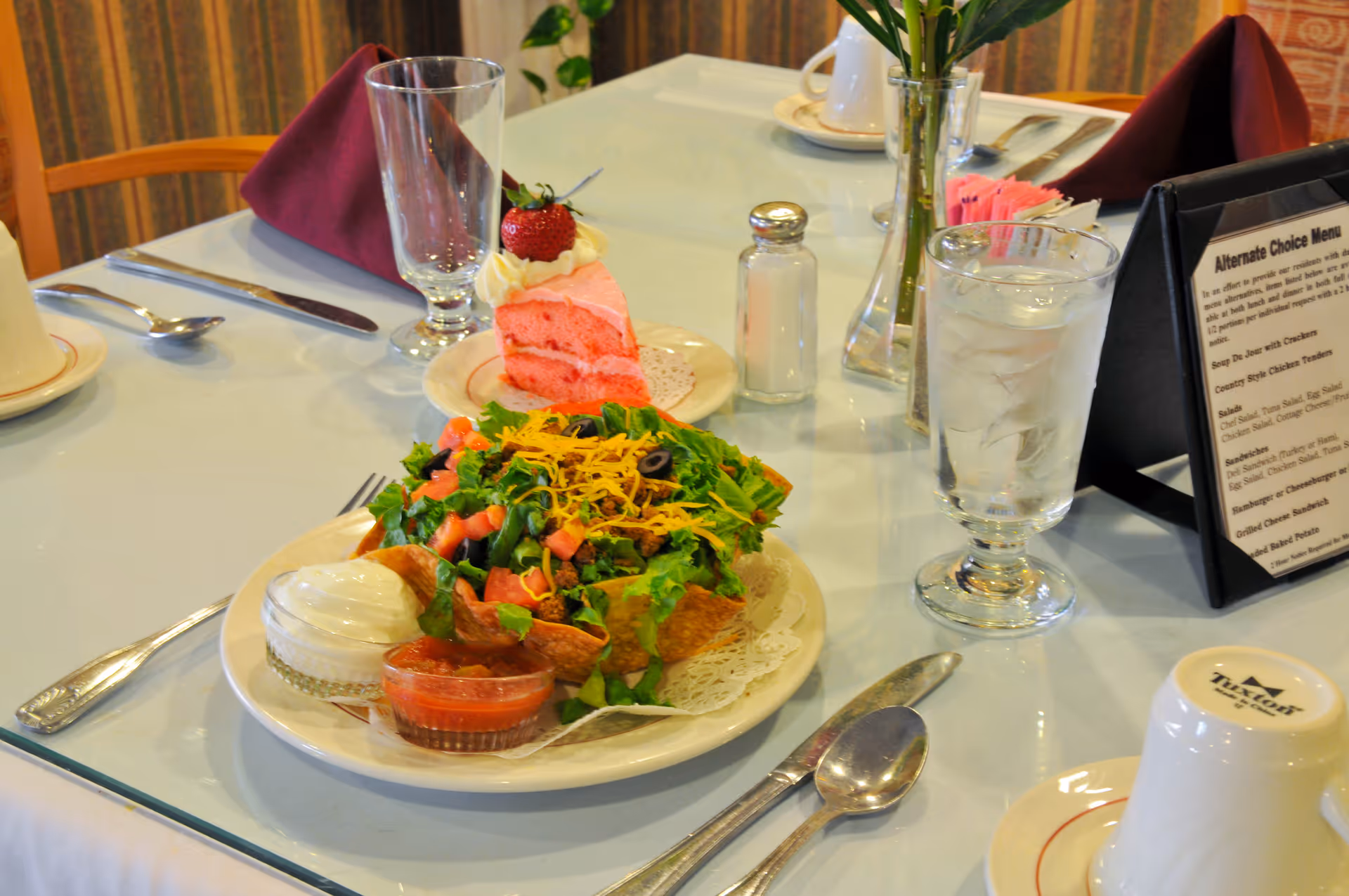 A dining table with a salad in a tortilla bowl, a pink slice of cake topped with a strawberry, a glass of water, and place settings.