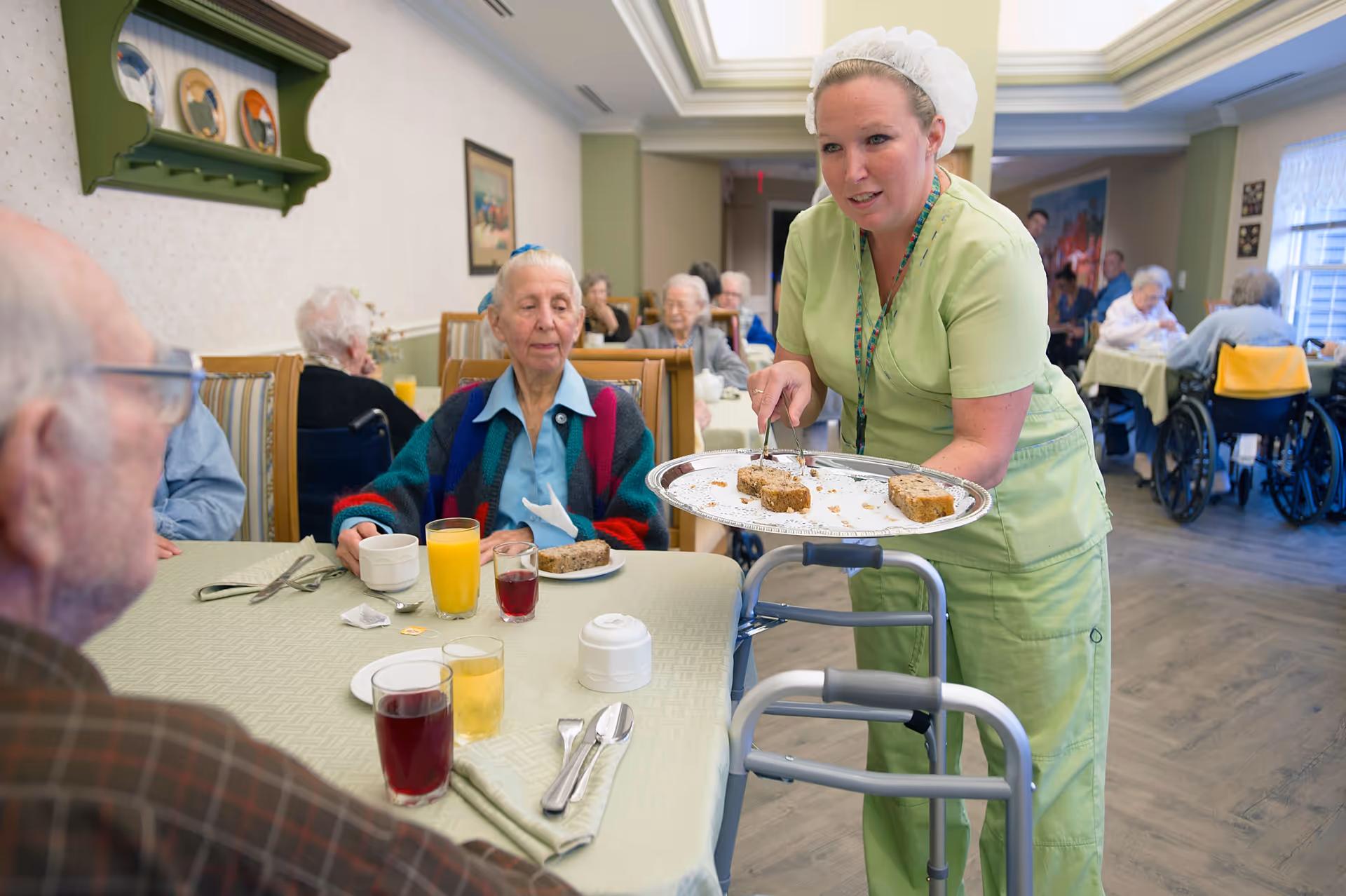 A caregiver in green scrubs and a white hairnet serves slices of cake to elderly residents seated at tables in a dining room of a senior living facility. The room has light green walls, framed pictures, and several elderly people sitting and eating or drinking.