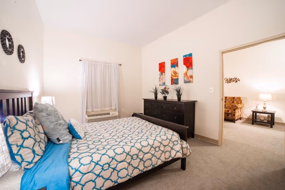 A bedroom with a bed featuring a blue and white patterned bedspread and multiple pillows. There is a dark wooden dresser with three decorative plants and three colorful floral paintings above it. A window with white curtains and an air conditioning unit is on the left wall. The room opens into a living area with a patterned armchair, a side table, and a lamp.