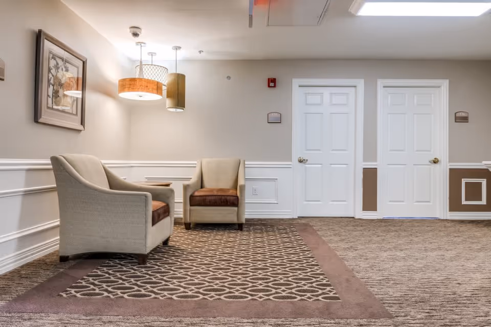 A seating area in a hallway with two beige armchairs with brown cushions placed on a patterned carpet. The walls are painted beige with white wainscoting and there are two white doors in the background. Three pendant lights hang from the ceiling and a framed picture is mounted on the wall.