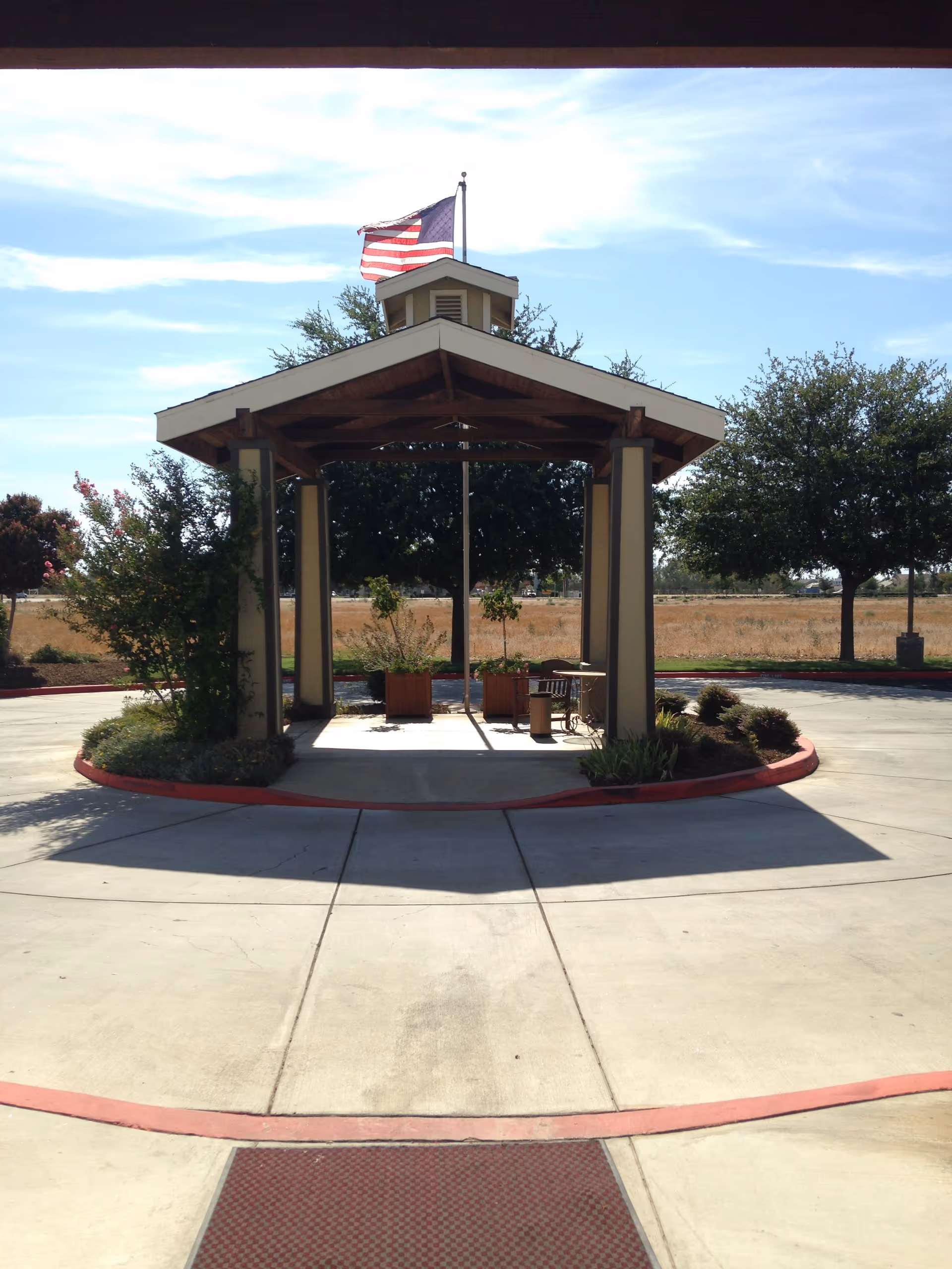 A small pavilion structure with a peaked roof and an American flag flying on top. The pavilion is surrounded by a circular concrete driveway with red curbing. There are some plants and trees around the pavilion and in the background, under a clear blue sky.