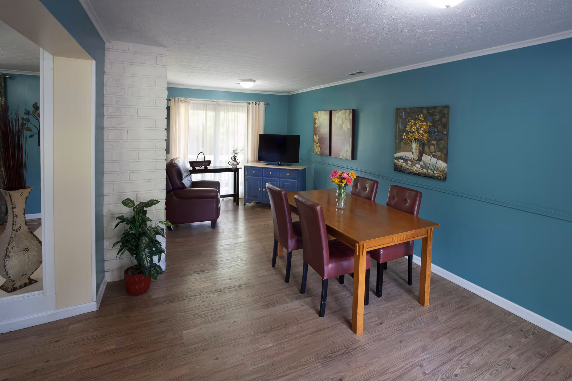 A cozy interior room with teal walls and wood flooring featuring a wooden dining table with four maroon chairs and a vase of colorful flowers on top. In the background, there is a brown leather armchair, a blue cabinet with a TV on it, and a window with sheer curtains letting in natural light. The room is decorated with paintings and a potted plant near a white brick column.