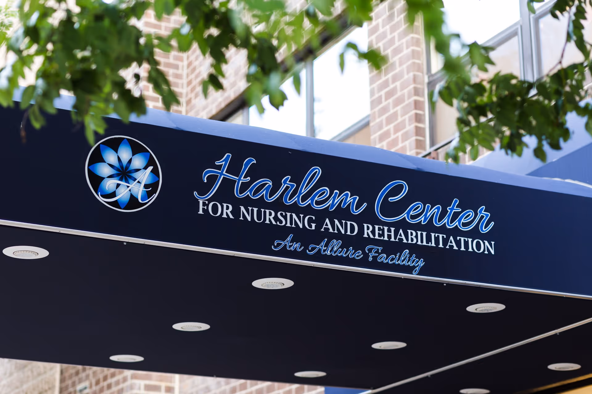 Blue entrance awning reading "Harlem Center for Nursing and Rehabilitation" with tree branches and a brick building facade in the background.
