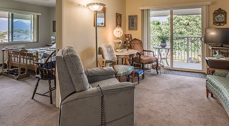 A cozy living area with a recliner chair, antique-style chairs, a small round table with a lamp, and a sliding glass door leading to a balcony with greenery outside. Adjacent to the living area is a bedroom with a wooden bed, a rocking chair, and a window showing a mountain view.
