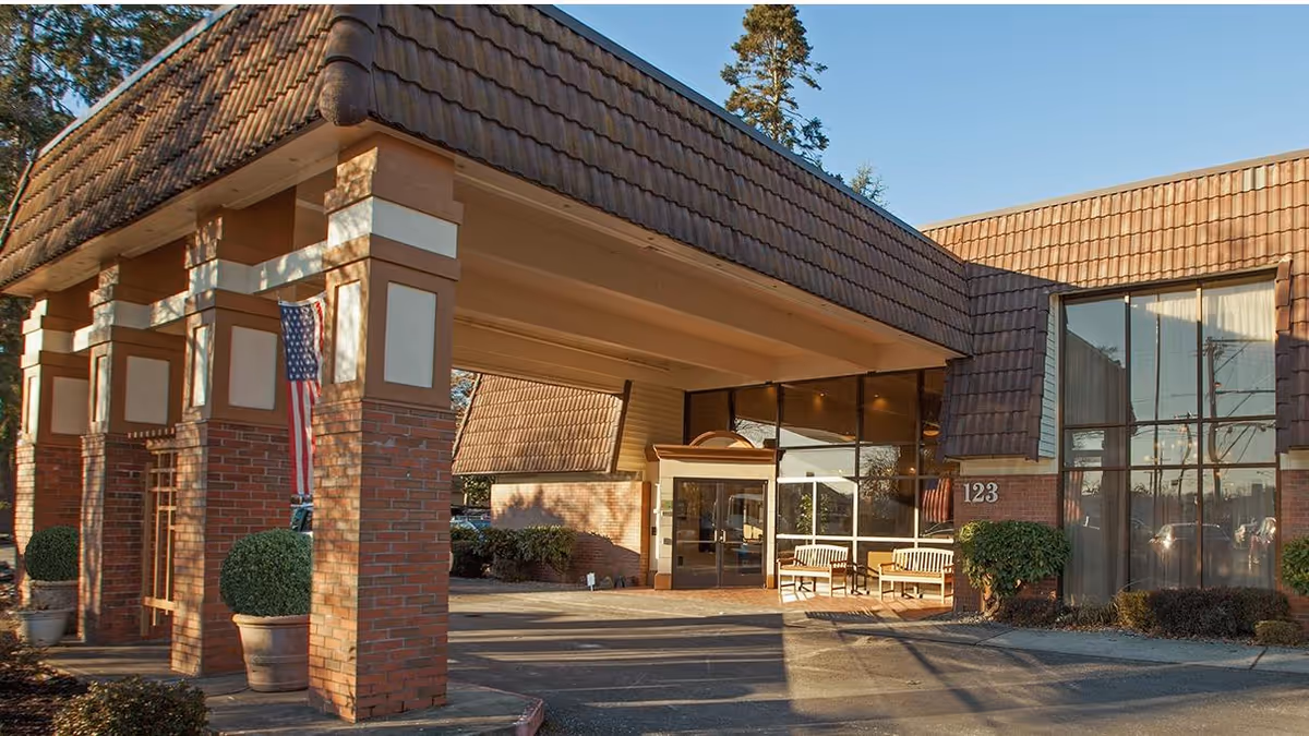Covered entrance and porte-cochere of a brick senior living building with benches, an American flag, and the number 123.