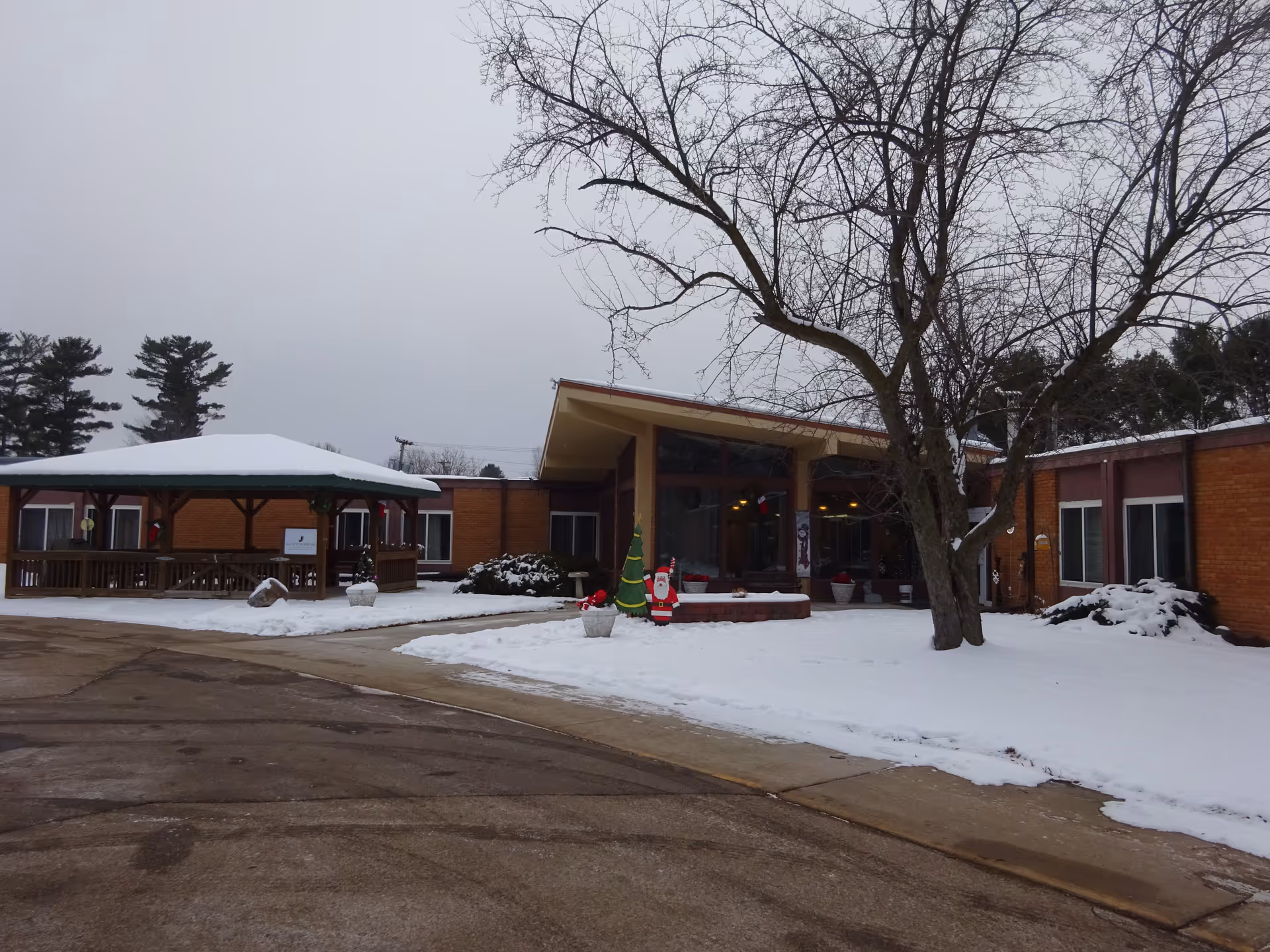 Snow-covered front entrance of a single-story brick senior living building with a leafless tree, a covered gazebo, and holiday decorations.