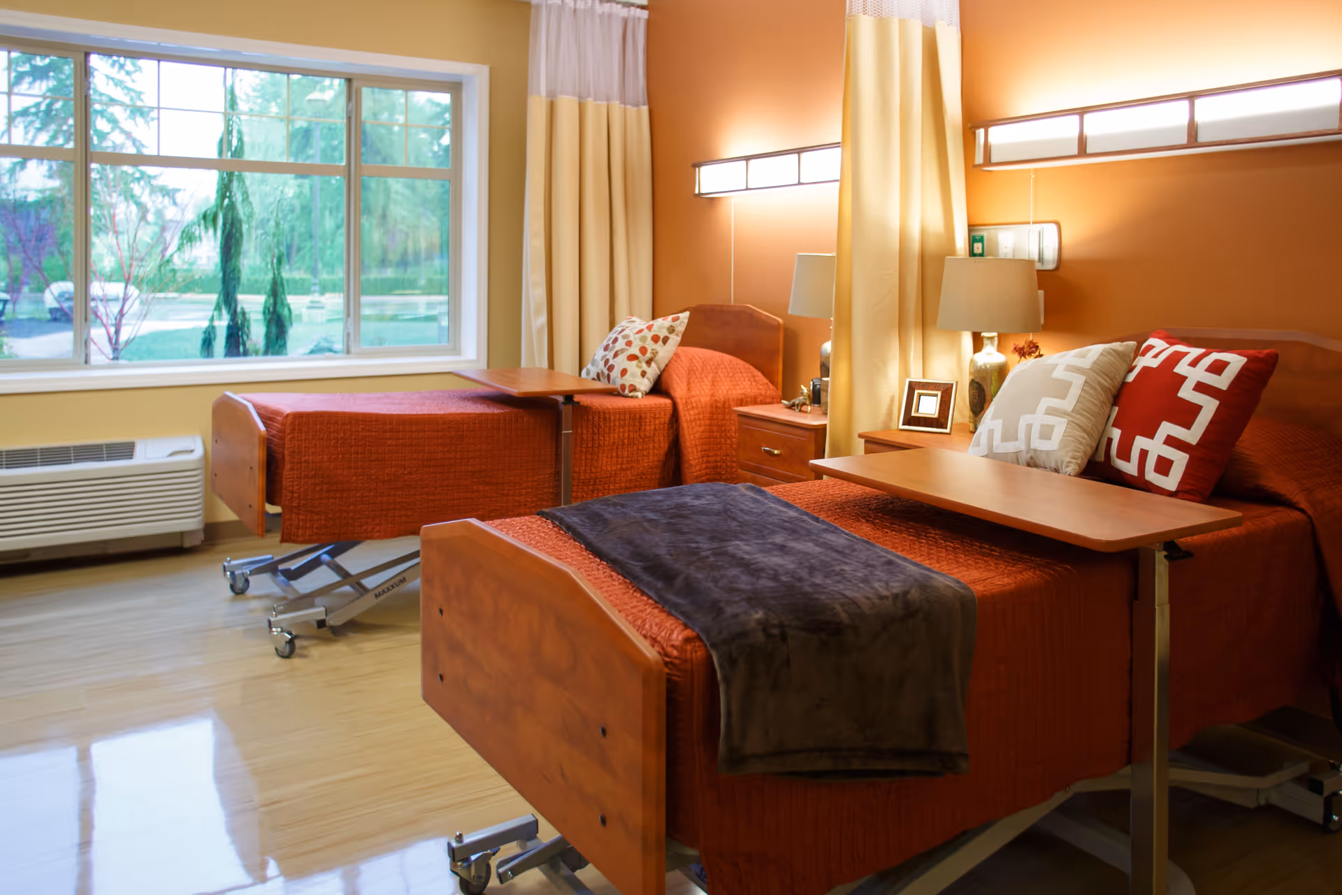 Two adjustable hospital-style beds with orange bedding, bedside tables, and a large window in a bright senior care room.