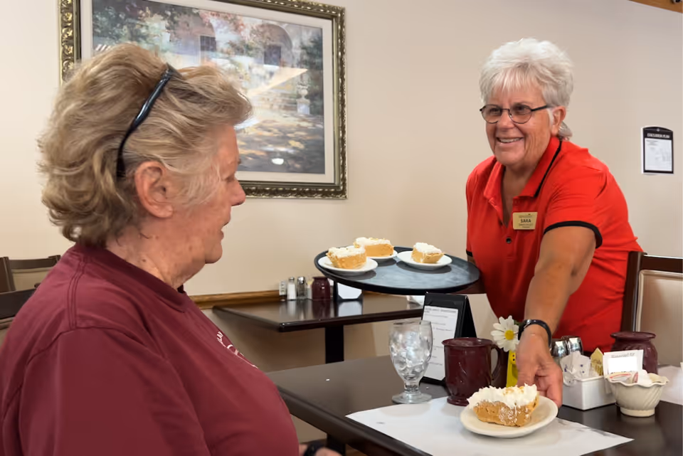 A smiling server in a red shirt hands a plate with dessert to an elderly woman seated at a dining table in a dining room.
