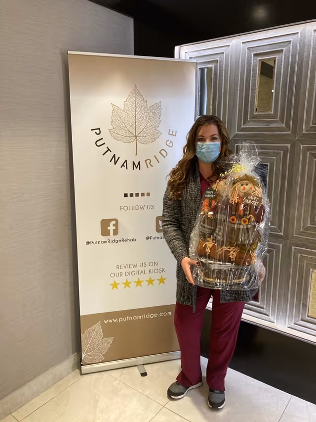 A woman wearing a face mask and maroon scrubs stands indoors holding a wrapped gift basket decorated with fall-themed items including scarecrows. Behind her is a banner with the Putnam Ridge logo, social media handles, and a website URL.