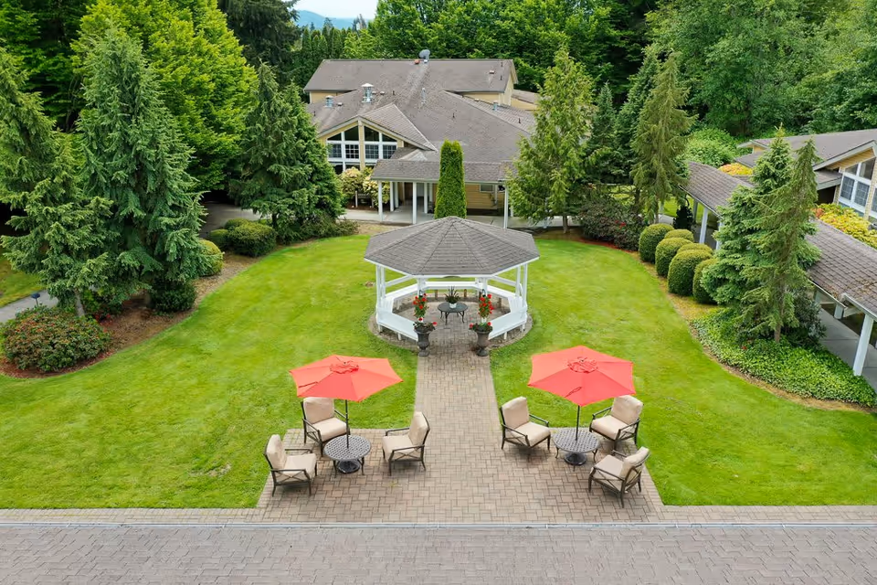 A well-maintained outdoor garden area at The Cottages of Snohomish featuring a central gazebo surrounded by green grass and trees. There are two seating areas with beige chairs and tables shaded by red umbrellas on a paved patio.