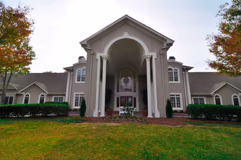 Front exterior view of a large senior living facility building with tall white columns at the entrance, surrounded by green grass and trees with autumn foliage.