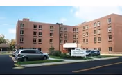 Exterior view of a multi-story brick building with several windows, identified as Kane Community Living Center Glen Hazel by a sign in front. There are a few parked cars and a clear sky above.