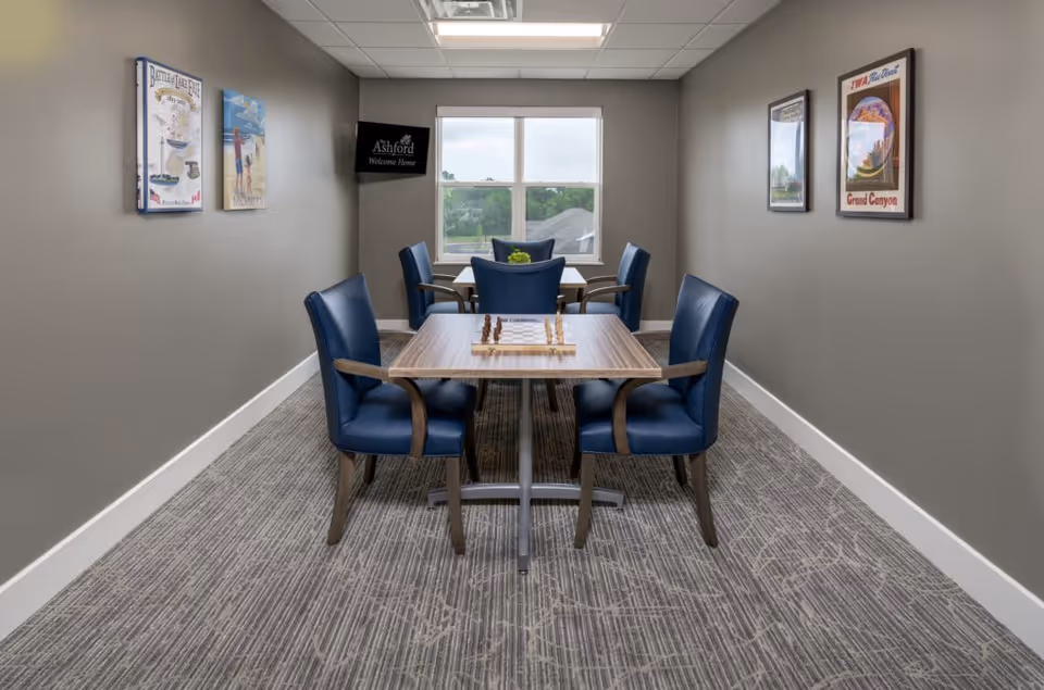 Small communal room with two square tables and blue chairs facing a window, a chessboard on the nearest table, and framed prints on gray walls.