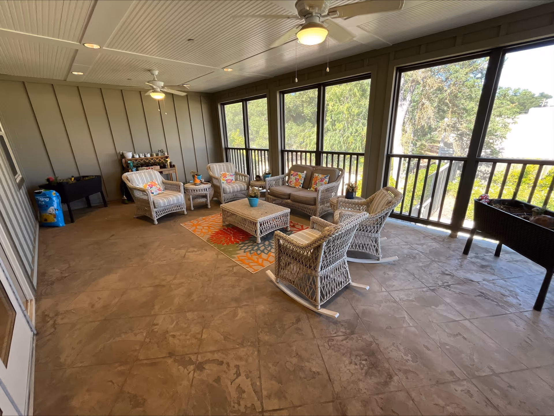 A spacious enclosed patio area with large windows overlooking greenery. The space features wicker furniture including a loveseat, four armchairs, and a coffee table on a colorful floral rug. Ceiling fans with lights are mounted on the white paneled ceiling. There is a small table with various items and a bag of potting soil in one corner.