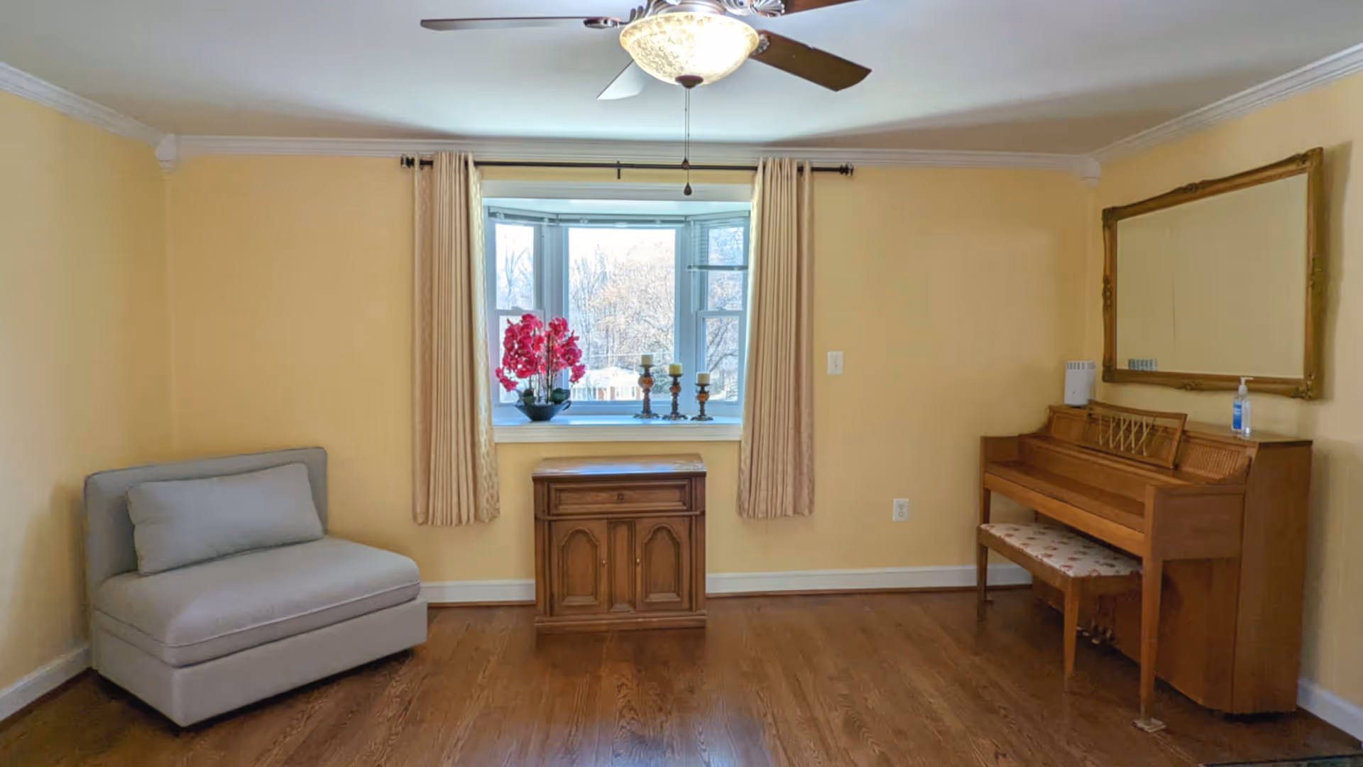 A cozy room with yellow walls and wooden flooring featuring a gray cushioned chair on the left, a wooden cabinet under a window with beige curtains, a vase with pink flowers, three candle holders on the windowsill, and a wooden piano with a floral cushioned bench on the right side beneath a large framed mirror.