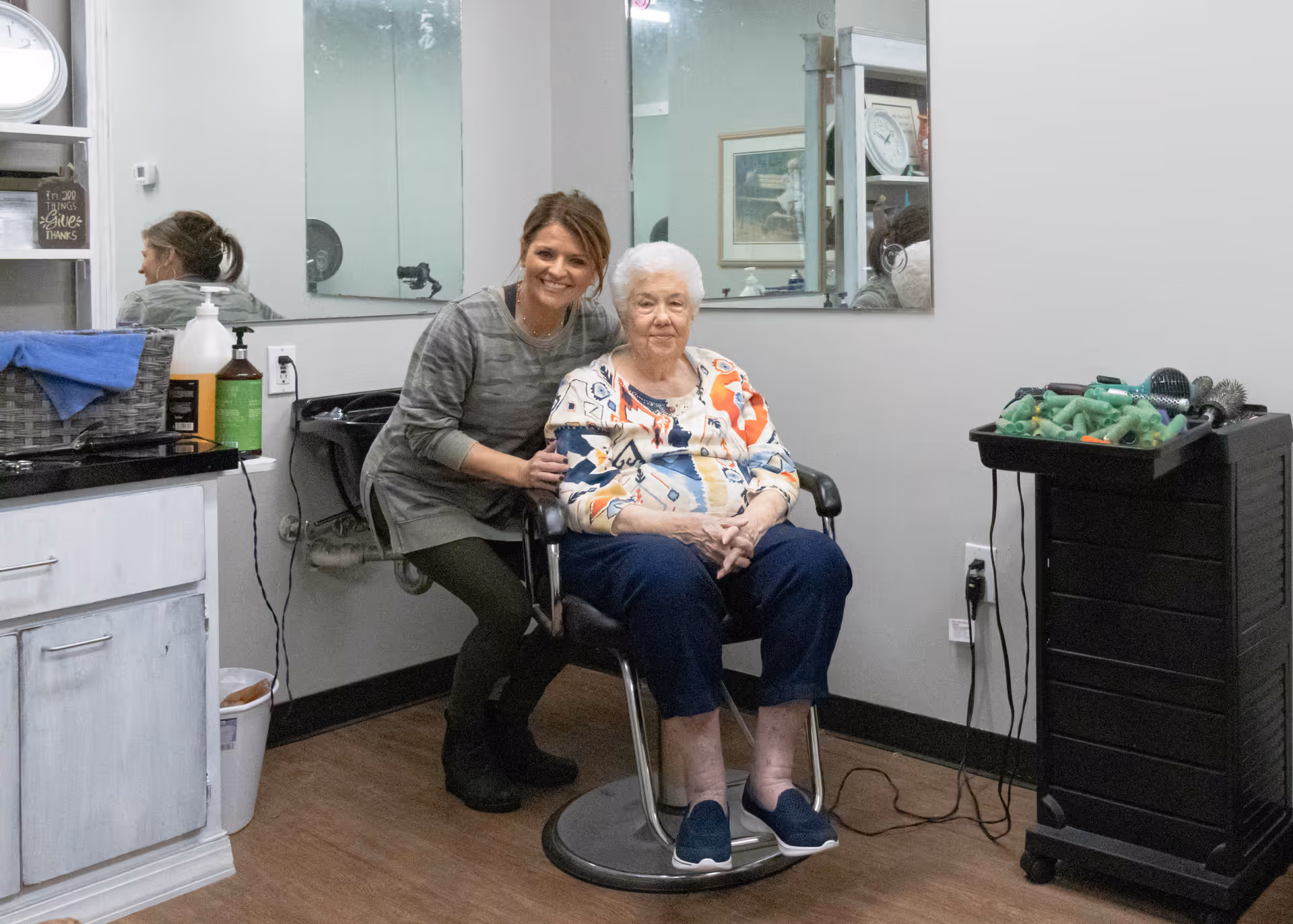 An elderly woman sitting in a salon chair with a younger woman standing beside her smiling. The room has a large mirror on the wall, a counter with hair care products, and a black rolling cart with hair styling tools.
