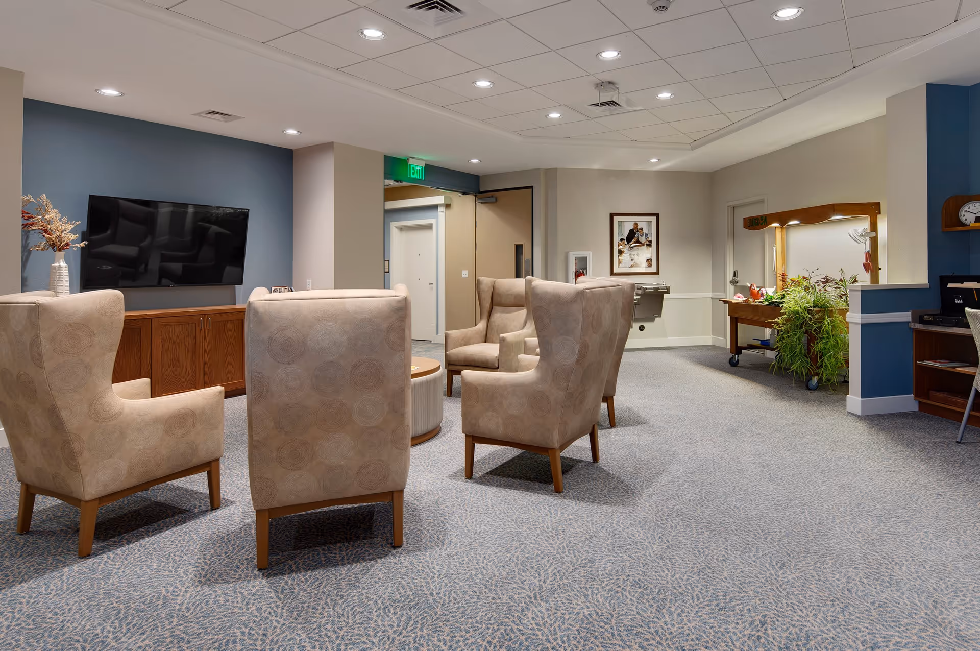 A cozy common area in a senior living facility with four beige upholstered armchairs arranged around a round wooden coffee table. A flat-screen TV is mounted on a blue accent wall above a wooden cabinet. The room has carpeted flooring, recessed ceiling lights, a water fountain, a framed picture on the wall, and a plant on a wooden cart near a doorway.