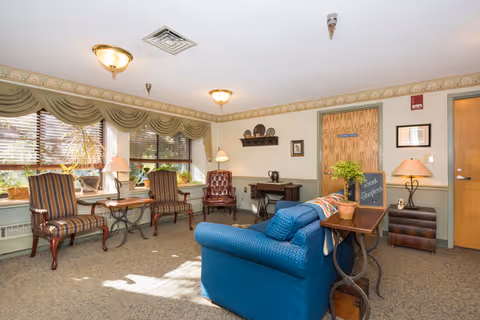 A cozy living room area with a blue upholstered sofa, two striped armchairs, and a burgundy leather chair. There are large windows with wooden blinds and green valances letting in natural light. The room has beige wallpaper with a floral border and carpeted floor. A small wooden table with a plant and a chalkboard sign that reads 'Good Shepherd' is placed behind the sofa. There are lamps, framed pictures, and decorative plates on the walls and shelves.