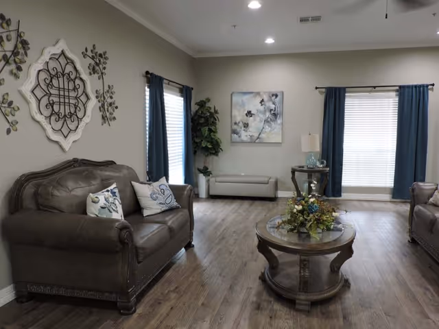 A cozy living room with two brown leather sofas facing each other, a round wooden coffee table with a floral arrangement in the center, a side table with a lamp, wall art, and two windows with blue curtains allowing natural light into the room.