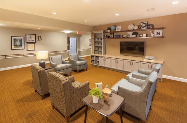 A cozy living room area in a senior living facility with several upholstered armchairs arranged around a small table with a plant and decorative items. The room features a large flat-screen TV mounted on a wall with built-in cabinets and shelves holding books, framed photos, and decorative objects. The carpet is brown, and the walls are light beige with framed artwork and a letter 'B' on one wall. The space is well-lit with recessed ceiling lights.