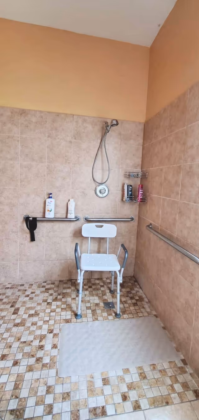 A tiled shower area with beige and brown mosaic floor tiles and light brown wall tiles. The shower has a handheld showerhead attached to the wall, a white shower chair with armrests, and grab bars on the walls for support. There are bottles of shampoo and body wash on a wall-mounted shelf and a grab bar with a black strap hanging from it. A non-slip mat is placed on the floor in front of the shower chair.