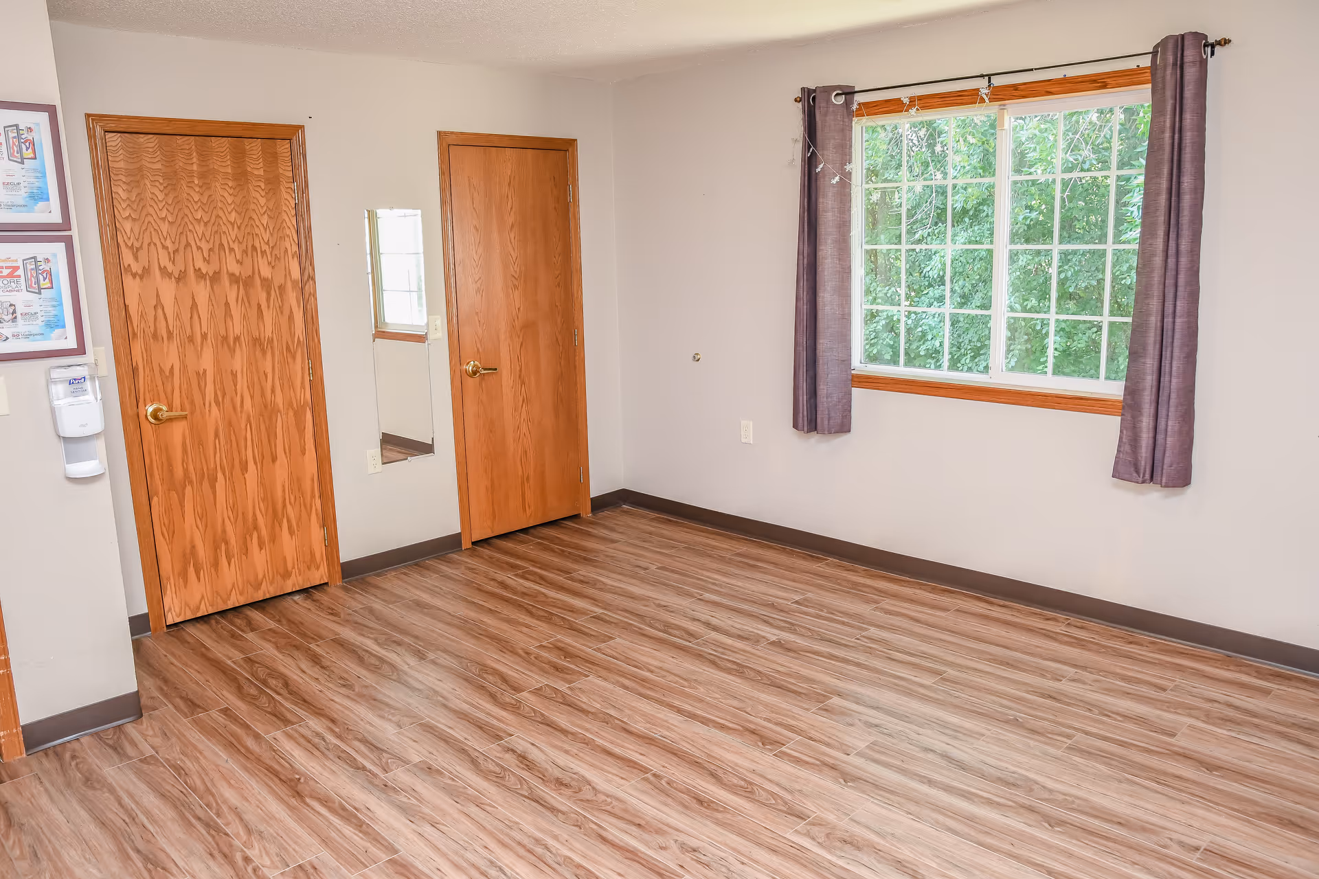 Empty room with wood-patterned vinyl flooring, two wooden doors with brass handles, a rectangular mirror on the wall between the doors, a large window with purple curtains showing green foliage outside, and light-colored walls.