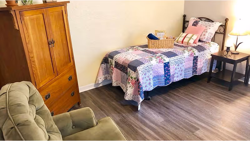 A cozy bedroom with a single bed covered in a patchwork quilt, a wooden cabinet, a green upholstered armchair, and a small side table with a lamp. There is a basket on the bed containing a book titled 'Word Hunt'. The room has wood flooring and light-colored walls.