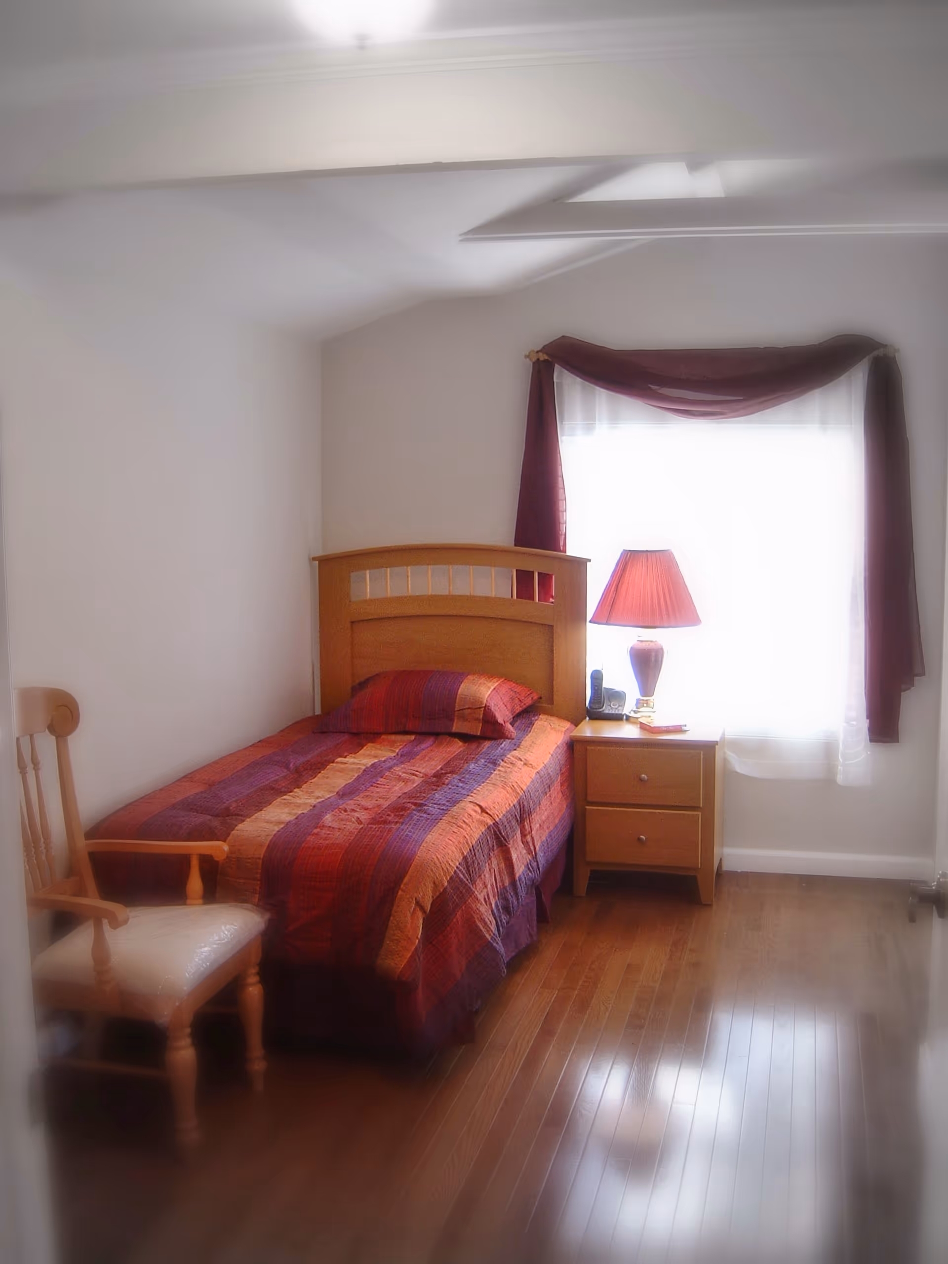 A small bedroom with a single bed covered in a striped quilt, a wooden nightstand with a lamp by a curtained window, and a chair.