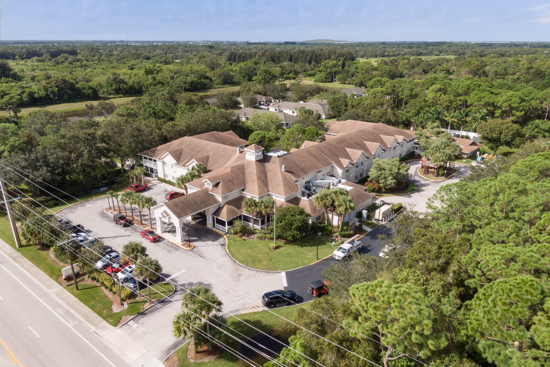 Aerial view of a senior living facility named Lake Forest Park surrounded by lush green trees and vegetation. The building has a brown roof and white exterior walls, with a parking lot featuring several cars. The facility is situated near a road and has landscaped areas with palm trees and other greenery.