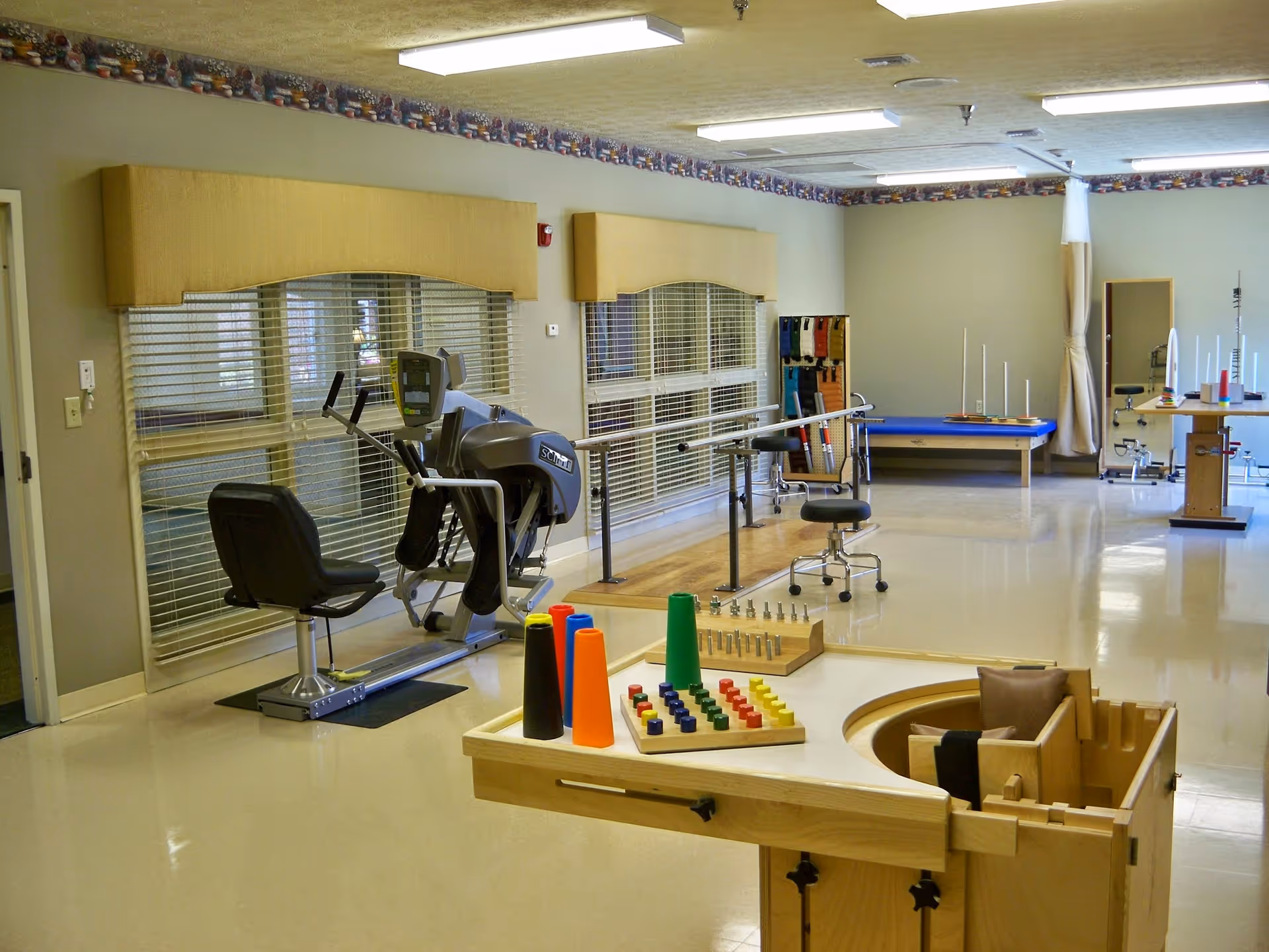 A spacious rehabilitation or therapy room with exercise equipment including a seated elliptical machine, parallel bars for walking support, a therapy table, and various colorful therapy tools and blocks on a wooden table. The room has large windows with blinds, a chair on wheels, and bright overhead lighting.