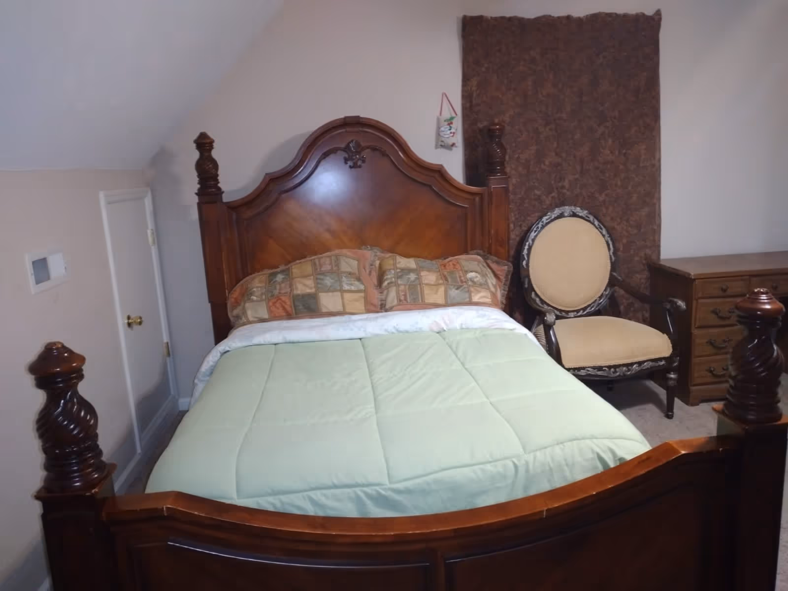 A bedroom with a wooden bed frame featuring ornate posts and a headboard. The bed is made with a light green comforter and two patterned pillows. To the right of the bed is a cushioned armchair with a wooden frame and a wooden dresser with drawers. A brown curtain hangs on the wall behind the chair.