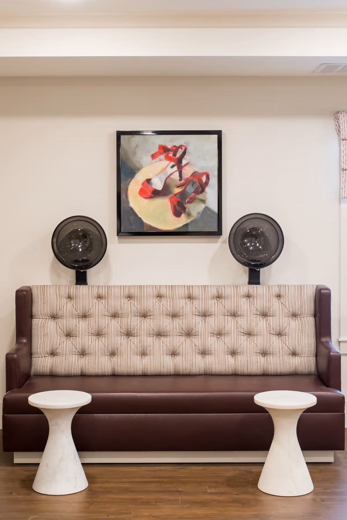 A salon area with a brown leather and fabric tufted bench seat flanked by two white marble side tables. Above the bench are two black hair dryer chairs and a framed painting of red high-heeled shoes on a round table.