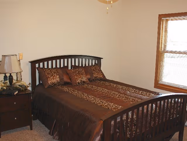 A neatly made bed with brown patterned bedding and matching pillows in a simple bedroom with a nightstand, lamp, and a window.