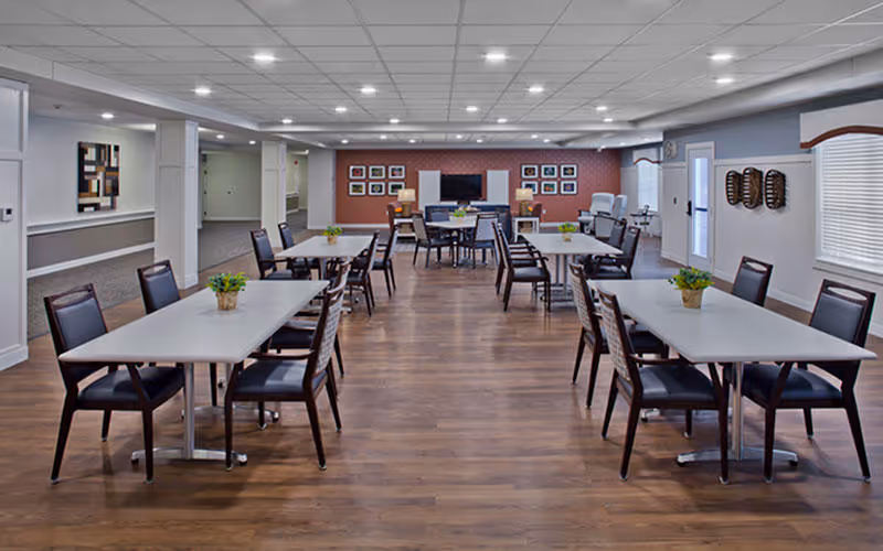 Spacious communal dining/activity room with multiple tables and chairs, wood flooring, a TV and framed pictures on the far wall.