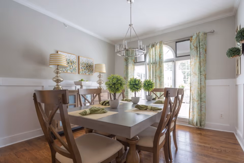 A bright dining room with a rectangular wooden table set for six, featuring green napkins and small potted plants as centerpieces. The room has wooden chairs with cushioned seats, two table lamps with striped shades on a sideboard, patterned curtains on large windows, and light gray walls with white wainscoting.