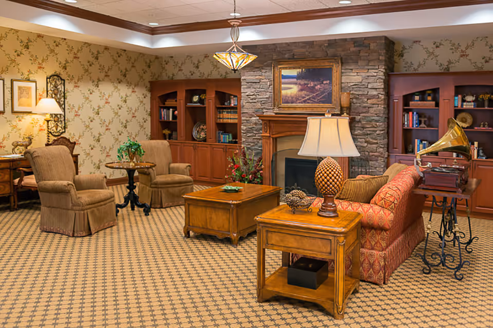 A cozy living room with patterned carpet, floral wallpaper, two upholstered armchairs around a small round table, a red patterned sofa, wooden coffee and side tables, a table lamp, a stone fireplace with a framed landscape painting above it, built-in wooden bookshelves filled with books and decorative items, and a vintage gramophone on a wrought iron stand.