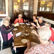 A group of elderly women sitting around a wooden dining table in a senior living facility, raising their glasses in a toast and smiling. The table is set with plates, glasses, and utensils, and there is a wine rack visible in the background.