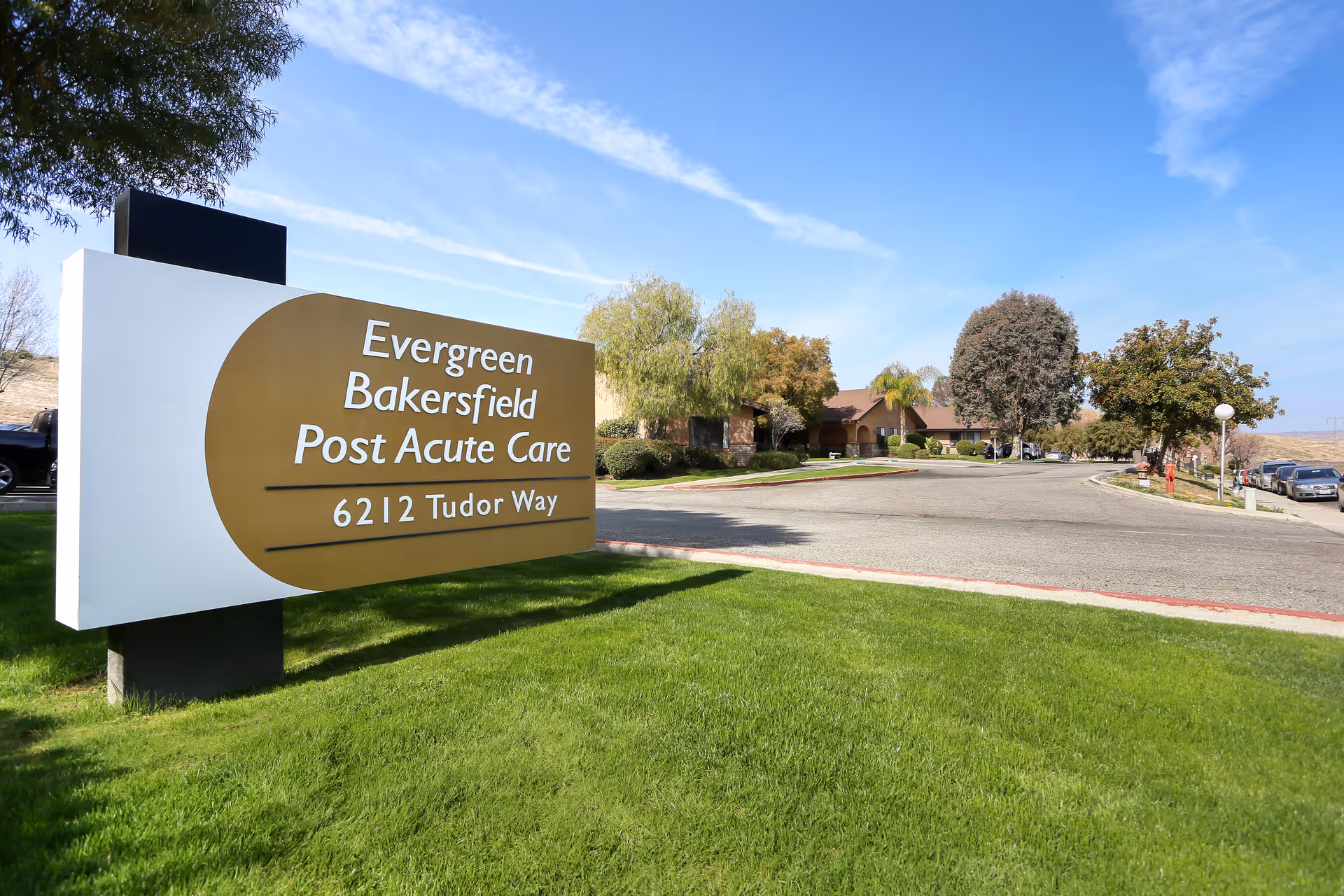 Outdoor view of the entrance area to Evergreen Bakersfield Post Acute Care facility, featuring a large sign with the facility name and address 6212 Tudor Way, surrounded by green grass, trees, and a driveway leading to the building under a blue sky.