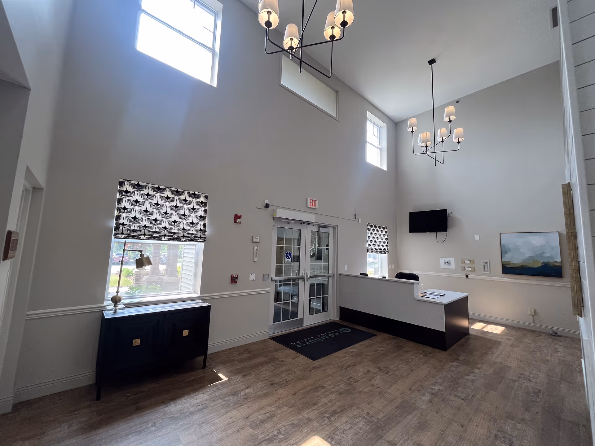 Interior view of a senior living facility reception area with high ceilings and large windows. The space features a reception desk with a chair, a wall-mounted TV, two windows with patterned blinds, a small cabinet with a lamp, and a painting on the wall. The floor is wooden, and there are two modern chandeliers hanging from the ceiling.