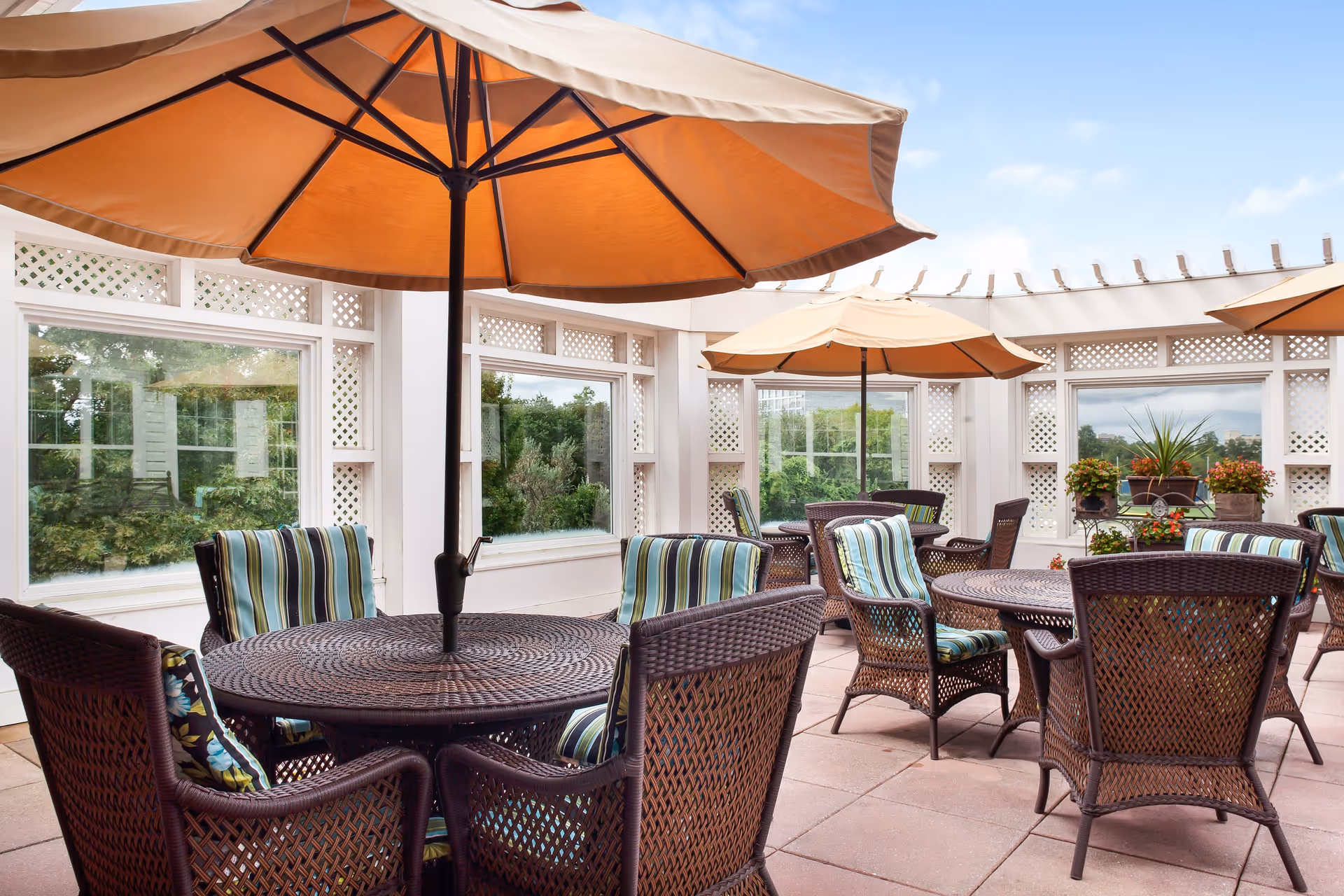 Sunny outdoor patio with wicker tables and chairs, striped cushions, potted plants, and large orange umbrellas.