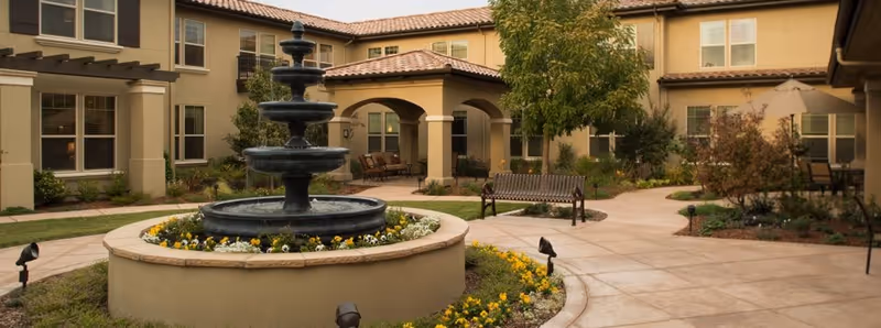 Outdoor courtyard area of a senior living facility with a multi-tiered black water fountain surrounded by a circular flower bed. The courtyard is paved with walkways and has benches, plants, and trees. The building surrounding the courtyard has beige walls, multiple windows, and a tiled roof.