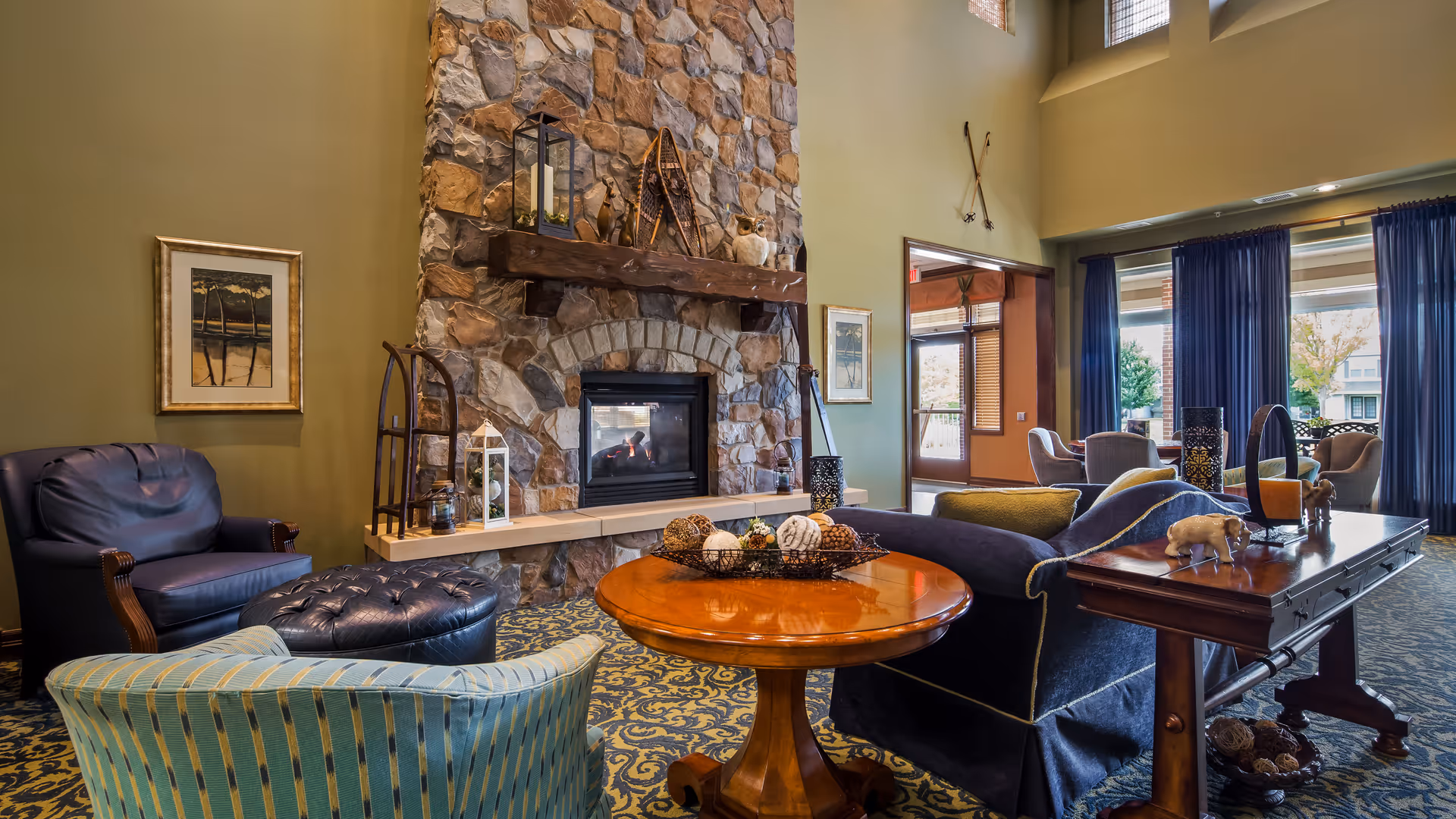 Cozy living room area with a stone fireplace, a wooden mantel decorated with lanterns and rustic items, a round wooden coffee table with decorative balls, a blue upholstered sofa, a striped armchair, and a leather armchair. Large windows with blue curtains allow natural light to fill the space.