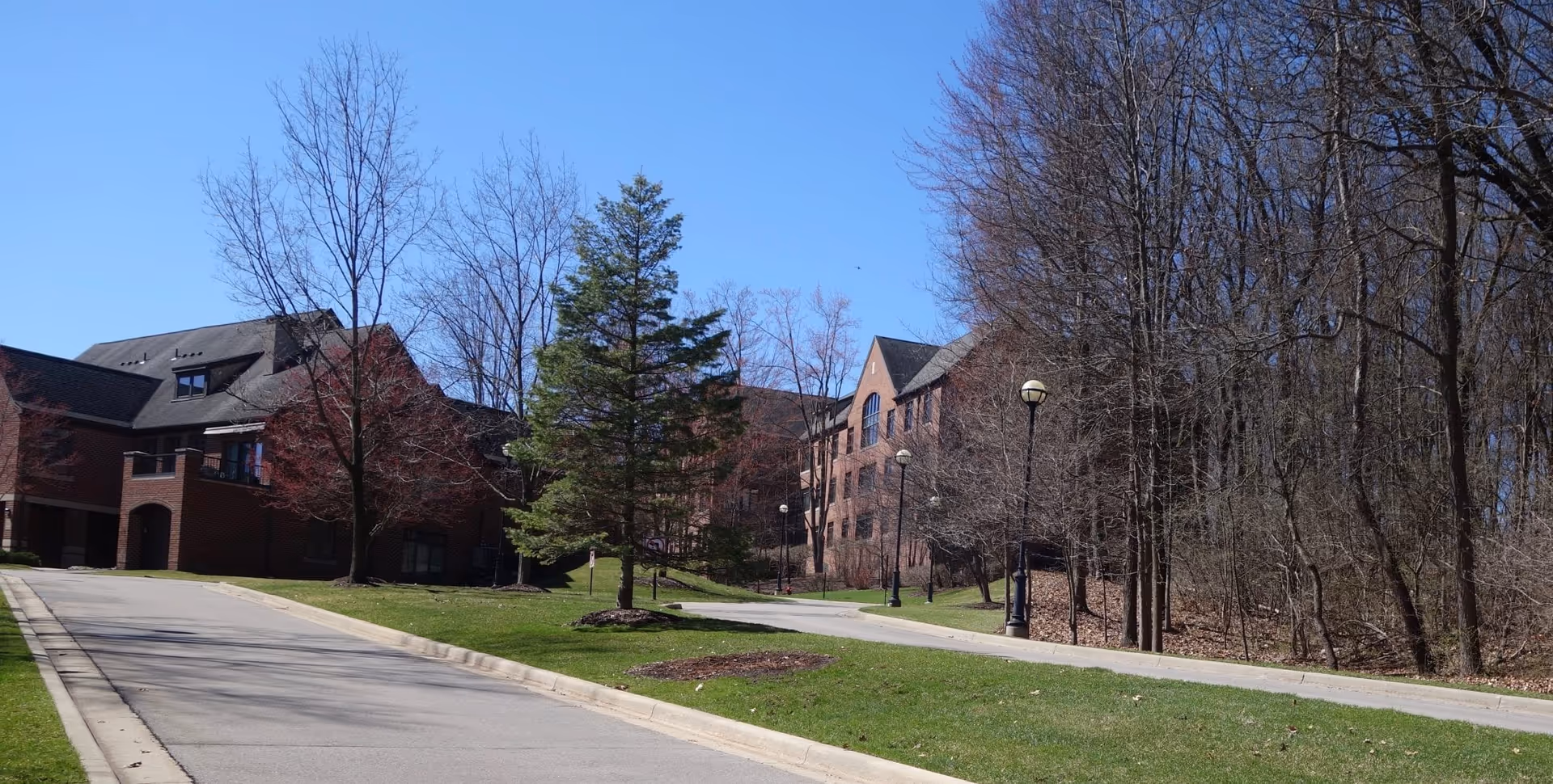 Brick senior living building with a curved driveway, lawn, trees, and lamp posts under a clear blue sky.