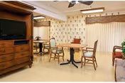 A common area in a senior living facility featuring a wooden TV cabinet with a television, a round table with four chairs, and a small dining table with two chairs. The room has patterned wallpaper, a ceiling fan, and vertical blinds covering a large window or sliding door.