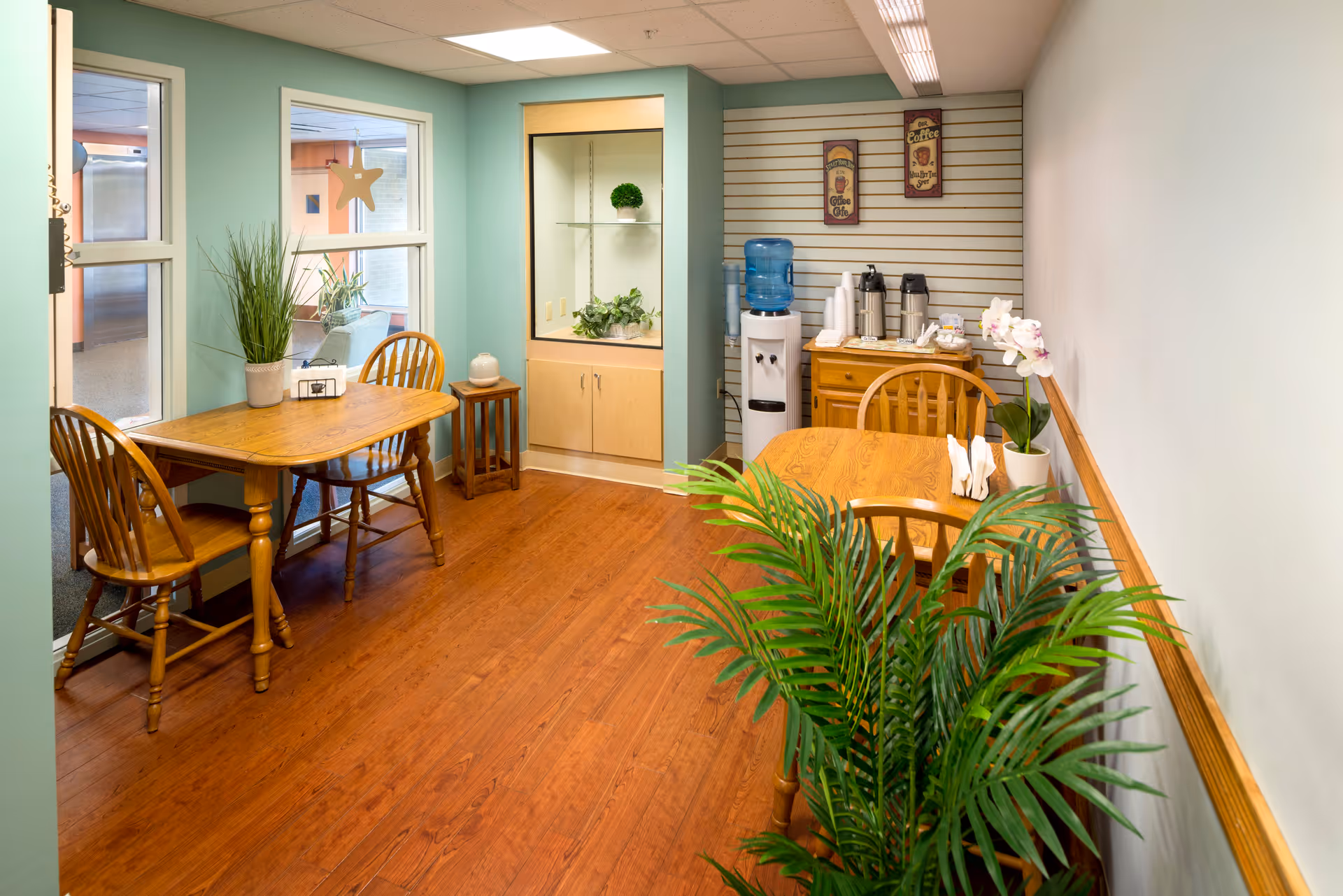 A small dining area with two wooden tables and chairs, a water dispenser, coffee station with cups and condiments, decorative plants, and wall art. The room has light blue and white walls and wood flooring.