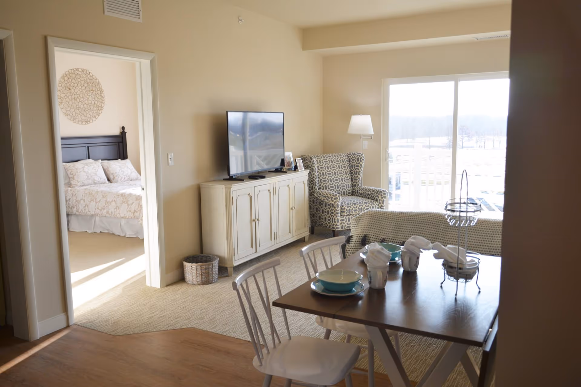 Interior view of a senior living facility showing a dining area with a wooden table set with plates, cups, and napkins. There are white chairs around the table. In the background, there is a living area with a patterned armchair, a TV on a white cabinet, and a large window letting in natural light. To the left, an open doorway reveals a bedroom with a bed and decorative wall art.