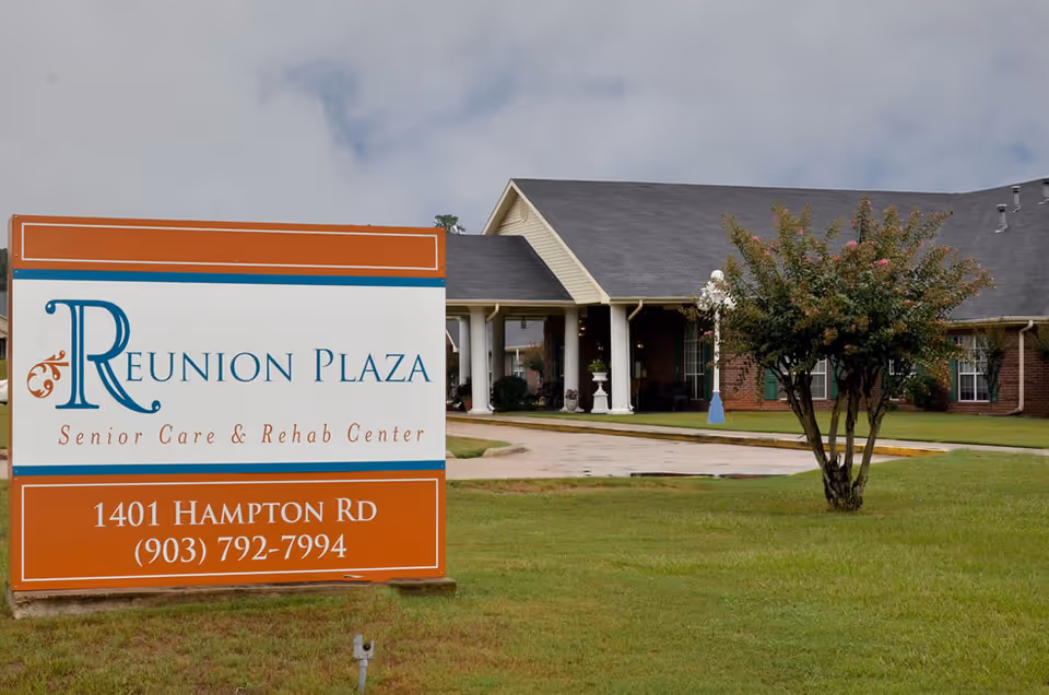 Large sign for Reunion Plaza Senior Care & Rehab Center in front of a single-story brick building with a covered entrance and a small tree on a grassy lawn.
